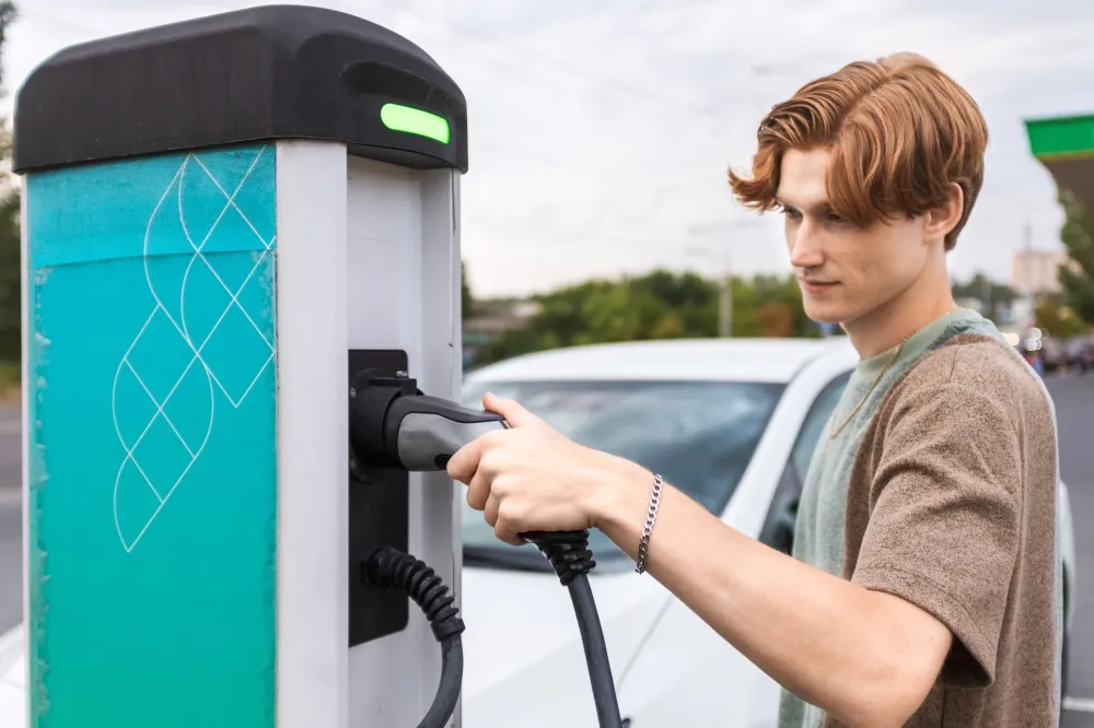Młody mężczyzna wyjmuje wtyczkę kabla z publicznej ładowarki samochodów elektrycznych, za nim stoi biały samochód.